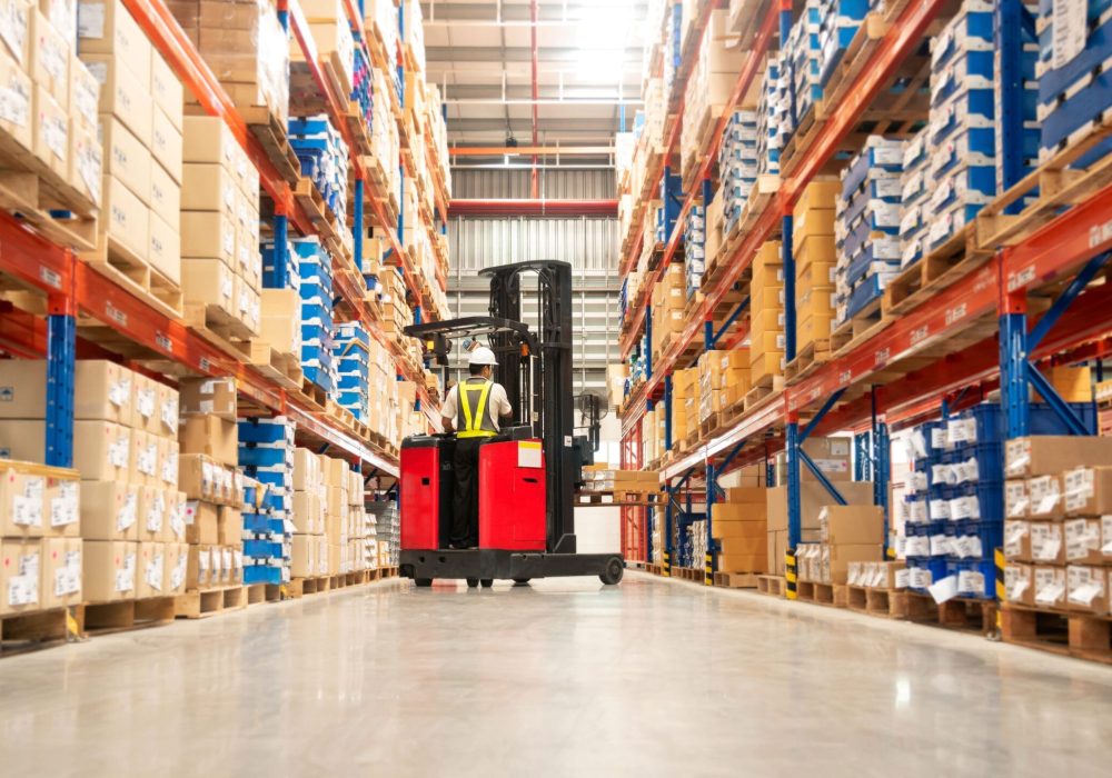 Worker in forklift-truck loading packed goods in huge distribution warehouse with high shelves.
